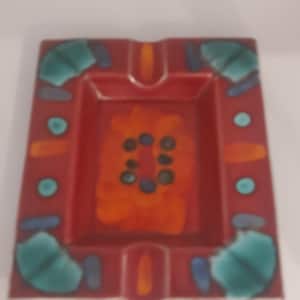 Könnte beinhalten: Quadratischer Keramik-Aschenbecher mit roter Basis und einem zentralen orangefarbenen Blumendesign. Der Aschenbecher hat türkisfarbene und blaue Akzente an den Rändern und in der Mitte. Rechteckige Form mit abgerundeten Ecken.