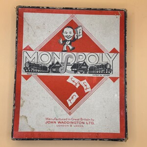 Op de afbeelding: Een vintage Monopoly bordspeldoos met een rood en wit ruitpatroon. De omslag bevat een stripfiguur met een pondteken, stoomtreinen en de tekst Monopoly, Manufactured in Great Britain by John Waddington Ltd. London & Leeds.
