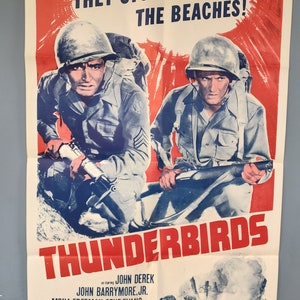 Może przedstawiać: Winietkowy plakat filmowy do filmu "Thunderbirds" z Johnem Derekiem. Plakat przedstawia czerwone tło z białą chmurą i tekstem "Szturmowali plaże!" Dwóch żołnierzy w mundurach wojskowych jest pokazanych na pierwszym planie, jeden trzyma karabin. Plakat jest klasycznym przykładem projektu plakatu filmowego wojennego.