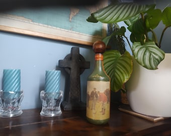 Puede incluir: Una botella verde con tapón de madera y una ilustración vintage de personas jugando al golf. Dos velas azules en espiral están en portavelas de cristal transparente. También se ven una cruz de madera oscura y una planta en maceta.