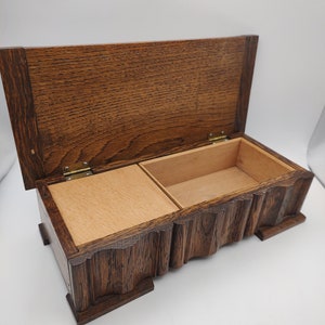 Op de afbeelding: Een houten doos met een scharnierend deksel en een gesneden ontwerp aan de voorkant. De doos is open en heeft een klein compartiment aan de binnenkant.