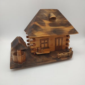 May include: A wooden model of a small house with a thatched roof, a well, and a fence. The house has a door and two windows. The model is painted in shades of brown and black.