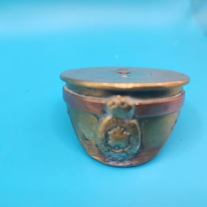 May include: A small, antique-style brass hat with a circular top and a decorative emblem on the front. The hat has a bronze and copper colour scheme, with a detailed emblem. The hat is set against a bright blue background.