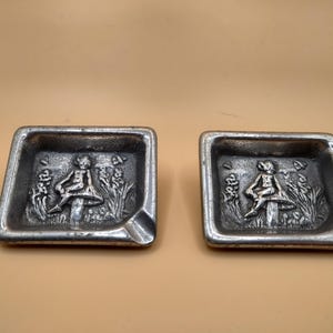 Puede incluir: Dos ceniceros de metal plateado con forma cuadrada y un diseño en relieve de un hada sentada en un hongo. Los ceniceros tienen un pequeño borde alrededor del borde.