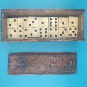 May include: A vintage domino set with ivory-coloured tiles and black dots, displayed in a wooden box. The box is open, revealing the dominoes, and the lid is placed below. The set is against a blue background.