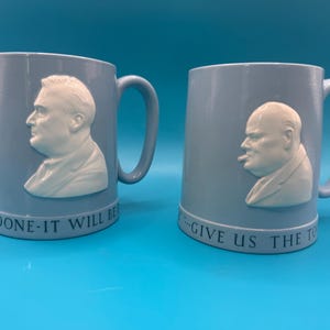 May include: Two light blue ceramic mugs featuring white relief portraits of historical figures. The left mug shows a profile of Franklin D. Roosevelt with the text BE DONE-IT WILL BE. The right mug shows Winston Churchill with the text GIVE US THE TO.