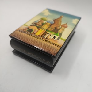 May include: A small wooden box with a black finish and a painted image of a colourful building with multiple domes. The box has a hinged lid and decorative gold trim.