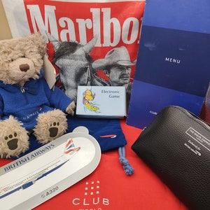 Puede incluir: Un oso de peluche rojo, blanco y azul que lleva una chaqueta azul con las palabras "British Airways" en ella. El oso está sentado sobre una superficie roja con una bolsa blanca detrás. La bolsa tiene la palabra "Marlboro" impresa en ella. También hay una pequeña bolsa de terciopelo azul con el logotipo de British Airways, un pequeño juego electrónico y una bolsa de tocador negra con las palabras "The White Company" y "Exclusively for British Airways" impresas en ella.