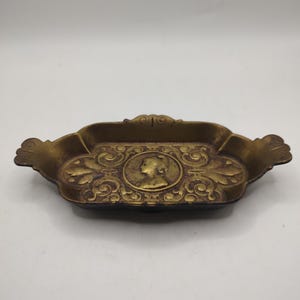 Puede incluir: Una pequeña bandeja de metal de color dorado con detalles ornamentados. La bandeja tiene un diseño ovalado en relieve en el centro que presenta el perfil de una mujer. La bandeja tiene dos asas a cada lado.