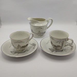 Puede incluir: Un juego de dos tazas de té blancas y platillos con un diseño plateado de un paisaje de montaña. También se incluye una jarra de crema blanca a juego con un diseño plateado de paisaje de montaña.