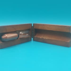 以下が含まれることがあります： 2 つに分かれた木製の型で、内部に丸みを帯びた円筒形の物体が入っています。型は温かみのある茶色で、物体も同様の素材でできているようです。背景は単色のティール色です。