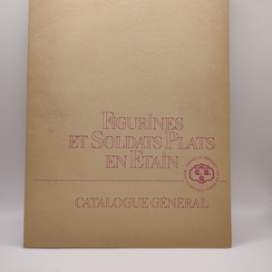 May include: A tan-colored catalog cover with the title "FIGURINES ET SOLDATS PLATS EN ETAIN" in red lettering. The cover also features a red circular logo with the text "MARQUE DE FABRIQUE" and "MOD&Egrave;LES D&Eacute;POS&Eacute;S FONDE EN 1911" in a smaller font. The text "CATALOGUE G&Eacute;N&Eacute;RAL" is printed in red at the bottom of the cover.