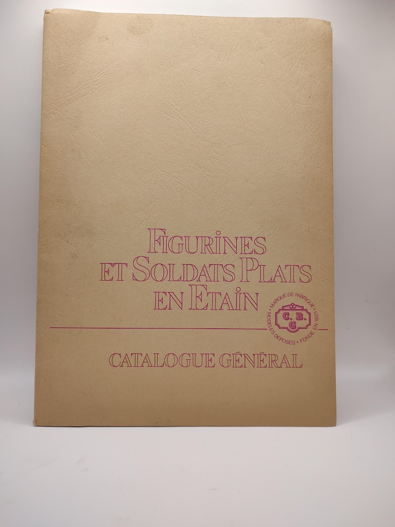 May include: A tan-colored catalog cover with the title "FIGURINES ET SOLDATS PLATS EN ETAIN" in red lettering. The cover also features a red circular logo with the text "MARQUE DE FABRIQUE" and "MOD&Egrave;LES D&Eacute;POS&Eacute;S FONDE EN 1911" in a smaller font. The text "CATALOGUE G&Eacute;N&Eacute;RAL" is printed in red at the bottom of the cover.