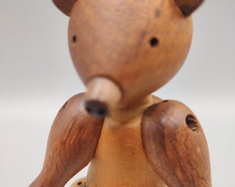 Puede incluir: Figura de oso de madera con cabeza redondeada, orejas pequeñas y extremidades articuladas. El oso está hecho de madera marrón clara y oscura, con vetas visibles. Los brazos y las piernas son articulados, lo que permite posarlo. Diseño sencillo y clásico.
