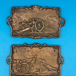 May include: Two rectangular bronze-toned metal plaques featuring embossed military scenes. The top plaque depicts soldiers with a cannon and the text UN VRAI POILU NOTRE 75 EN ACTION. The bottom plaque shows soldiers in a field with the text EN ALSACE LES DIABLES BLEUS.