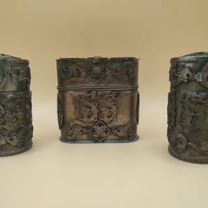 Puede incluir: Tres recipientes de plata con intrincados diseños de dragones. Los recipientes son de forma cilíndrica y rectangular. Los recipientes están hechos de plata y tienen un acabado deslustrado.
