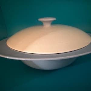 Op de afbeelding: Een lichtblauwe keramische serveerschaal met een brede, platte rand en een bijpassend koepelvormig deksel in een crèmewitte kleur. Het deksel is voorzien van een centrale knopgreep en de schaal heeft een gladde, afgeronde onderkant.