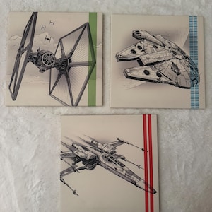 May include: Three square wall art panels featuring black and white ink-style illustrations of Star Wars spacecraft. One panel shows a TIE fighter with a green stripe, another the Millennium Falcon with a blue stripe, and the third an X-wing with red stripes.