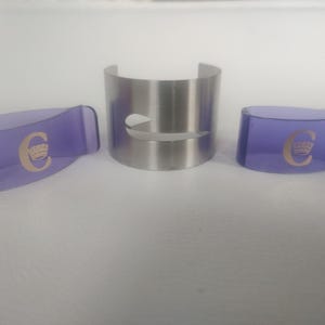 Puede incluir: Tres accesorios de bar: un brazalete de metal plateado con un recorte curvo y dos clips de plástico morado. Los clips tienen un logotipo "C" dorado con una corona. Los artículos están sobre una superficie blanca.