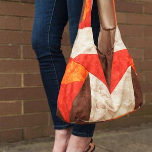 Könnte beinhalten: Eine bunte Tragetasche mit geometrischem Muster in Rot-, Orange-, Braun- und Cremetönen. Die Tasche wird von einer Person getragen, die eine dunkelblaue Jeans und braune High Heels trägt.