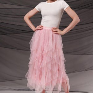 Op de afbeelding: Een vrouw draagt een witte top met korte mouwen en een roze tule rok met meerdere lagen. De rok heeft een gelaagd ontwerp. Zilveren sandaaltjes met hakken maken de outfit compleet. De achtergrond is een donkergrijze stof.