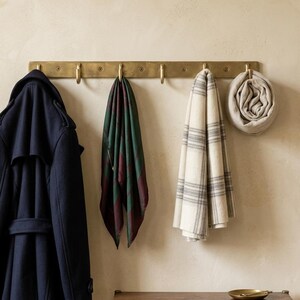 Peut inclure: Un porte-manteau mural horizontal en laiton doté de six crochets régulièrement espacés. Sont suspendus aux crochets un trench-coat bleu marine, une écharpe à carreaux vert et bordeaux, une écharpe à carreaux crème et gris, et une écharpe beige roulée.