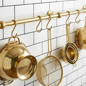 Peut inclure: Une barre de cuisine horizontale en laiton fixée au mur, équipée de plusieurs crochets en forme de S. Suspendus aux crochets se trouvent une grande passoire en laiton, une passoire fine ronde, des tasses à mesurer en laiton et une petite passoire.