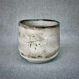 Peut inclure: Une tasse en céramique faite à la main avec un glaçage blanc cassé moucheté. La tasse a une forme arrondie avec un bord vert foncé et une ouverture légèrement évasée. L'extérieur présente des mouchetures marron foncé et vertes.