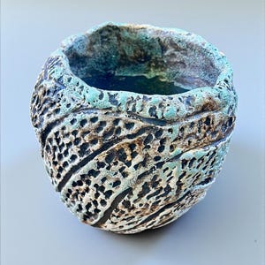 Peut inclure: Un bol en céramique fait à la main avec une surface texturée. Le bol présente un design unique avec une combinaison de couleurs noir, blanc et turquoise. Le bord est émaillé turquoise et l'extérieur présente un motif de trous et de lignes.