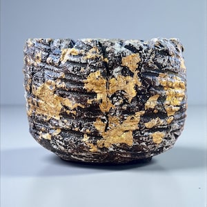 Peut inclure: Un bol en céramique texturé, avec une base marron foncé et des accents de feuilles d'or. Le bol présente un aspect brut et organique, avec des rainures visibles et un bord légèrement irrégulier. La feuille d'or ajoute une touche de luxe au design rustique.