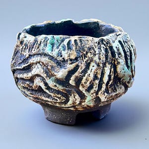 Pode incluir: Uma pequena tigela de cerâmica artesanal com um design texturizado e orgânico. A tigela apresenta uma base escura e terrosa e um exterior áspero e em camadas com tons de preto, branco e verde-azulado. A borda é escura e a tigela repousa sobre três pequenos pés.