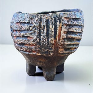 May include: A handcrafted, textured ceramic planter with a dark, earthy colour palette. The small, three-legged pot features a rough, ridged exterior with hints of blue, orange, and black. The open top is slightly irregular.
