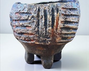 May include: A handcrafted, textured ceramic planter with a dark, earthy color palette. The small, three-legged pot features a rough, ridged exterior with hints of blue, orange, and black. The open top is slightly irregular.