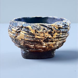 Puede incluir: Un cuenco de cerámica hecho a mano con un diseño texturizado y en capas. El cuenco presenta un interior azul oscuro y un exterior rugoso, dorado y marrón. La base es oscura y cuadrada. El cuenco es adecuado para servir o exhibir.