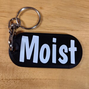 May include: Black rectangular keychain with the word "Moist" in white block letters. The keychain has a silver-colored metal ring and clasp for attaching to keys or a bag. The background is a light brown wooden surface.