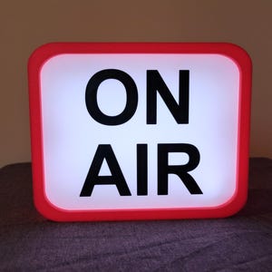 May include: A red rectangular sign with a white background and black text that reads "ON AIR".