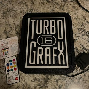 May include: Black sign with white text that reads "TURBO 16 GRAFX". The sign is illuminated with LED lights and comes with a remote control to change the color of the lights.