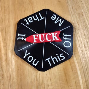 May include: A black, hexagonal novelty item with white text and a red arrow. The word "FUCK" is in the center, with phrases like "Me" and "Off" around the edges. The item is on a wooden surface.