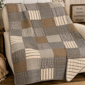 May include: A patchwork quilt featuring a repeating pattern of square fabric blocks in shades of gray, tan, and cream. The design incorporates solid colors, thin stripes, and small checkered prints, all stitched together with a visible quilted texture.