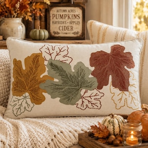 May include: A rectangular cream-colored throw pillow featuring embroidered autumn leaves in shades of mustard yellow, sage green, and burnt orange. The design includes detailed stitching on the leaves, with some outlined in thread against the fabric.