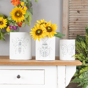 May include: Three white cylindrical vases with insect designs: a bee, a ladybug, and a butterfly. The ladybug vase holds yellow sunflowers. The vases are on a wooden shelf, with a white cabinet below.