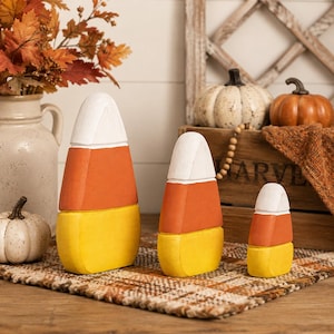 May include: Three tiered candy corn decorations in descending sizes, each featuring a white top, orange middle, and yellow base. These smooth, solid-colored objects are arranged in a row, showcasing a classic triangular shape with rounded corners.