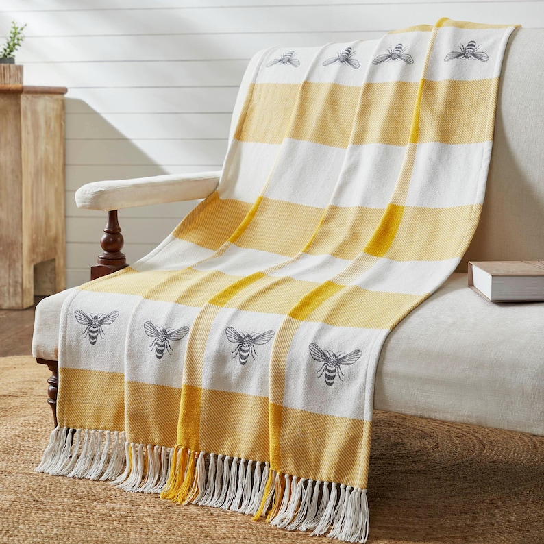 May include: A woven throw blanket featuring a repeating pattern of dark gray bees on alternating horizontal stripes of white and golden yellow. The blanket has a textured fabric finish and is finished with long, white fringe along the bottom edge.
