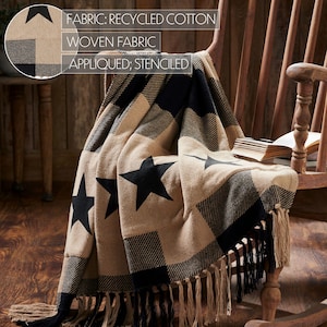 May include: A woven throw blanket featuring a black and tan buffalo check pattern with large black stenciled stars. The fabric is recycled cotton. The blanket has long, knotted fringe trim along the bottom edge. Text reads: FABRIC: RECYCLED COTTON, WOVEN FABRIC, APPLIED; STENCILED.