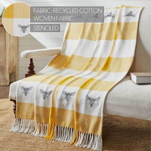 May include: A woven throw blanket featuring a yellow and white checkered pattern with stenciled bee motifs. The fabric is made of recycled cotton and includes a fringe trim along the bottom edge. Text on the image reads: FABRIC: RECYCLED COTTON WOVEN FABRIC STENCILED.