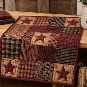 May include: A square quilted table mat featuring a patchwork design of burgundy, tan, and black plaid fabric squares. The mat is decorated with four dark burgundy fabric stars appliqued onto the corners and center, finished with a burgundy border.