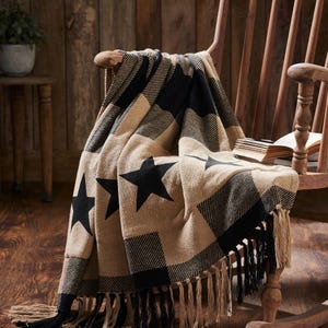 May include: A woven throw blanket featuring a black and tan buffalo check pattern with large black star motifs. The blanket has a textured knit construction and is finished with long, alternating black and tan fringe along the bottom edge.