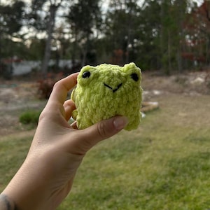 May include: A small, lime green crocheted frog with black eyes and a stitched mouth. The frog is made of a soft, textured yarn and has a rounded shape. The frog is a handmade item.
