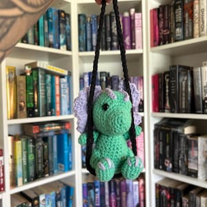 May include: A handmade crochet dinosaur toy in shades of green and purple, suspended by black cords. The dinosaur is held up against a backdrop of white bookshelves filled with books. The toy is a charming, whimsical creation.