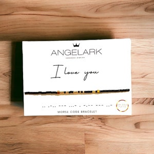 May include: A white box with the words "ANGELARK HANDMADE JEWELRY" and "I love you" in black text. A black beaded bracelet with gold accents is displayed on the box. The bracelet is a Morse code bracelet with the text "I love you" in Morse code. The box also has the text "MORSE CODE BRACELET" and "14K GOLD PLATED" in black text.